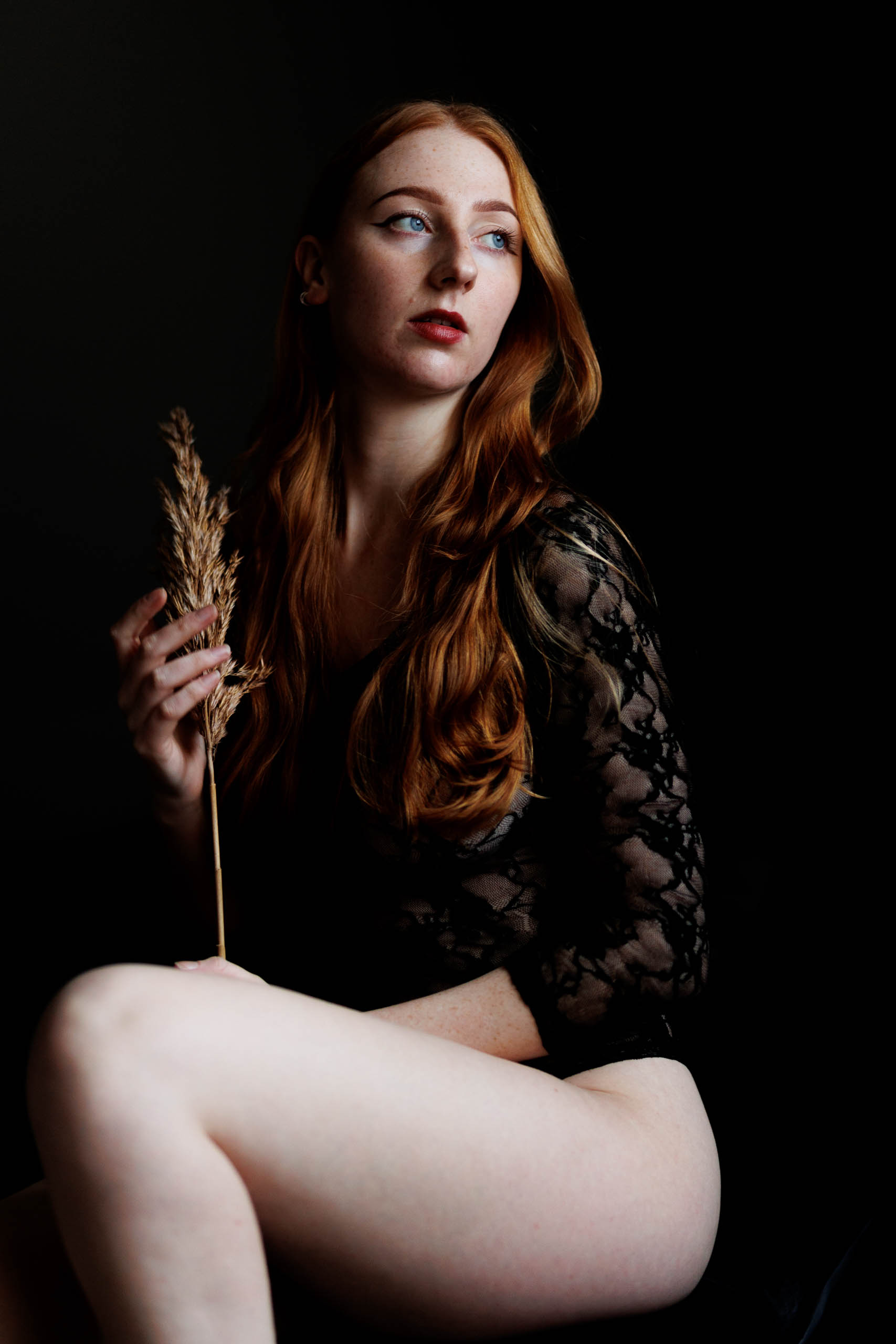 A woman with red hair sitting on a chair during a boudoir photo shoot in Rotterdam, elegantly holding a blade of grass.