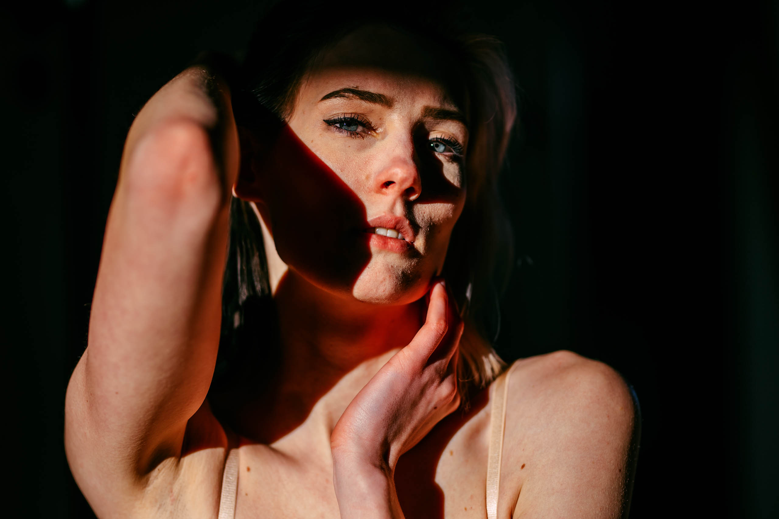 Een vrouw poseert in een donkere kamer tijdens een boudoirshoot, met haar hand op haar gezicht.