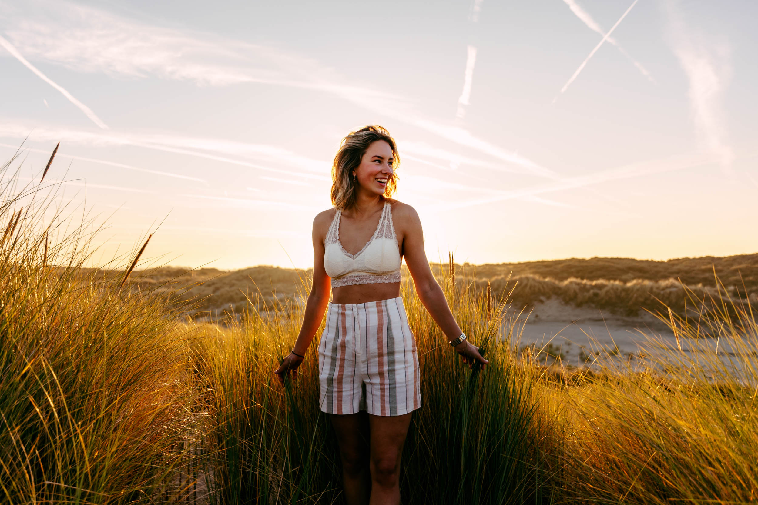 A woman poses for beach photos in tall grass at sunset.