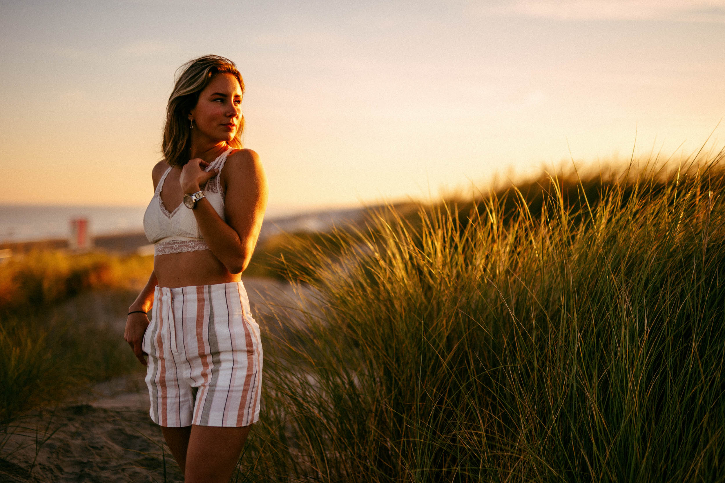 A woman in striped shorts standing on a beach at sunset, taking beach photos.