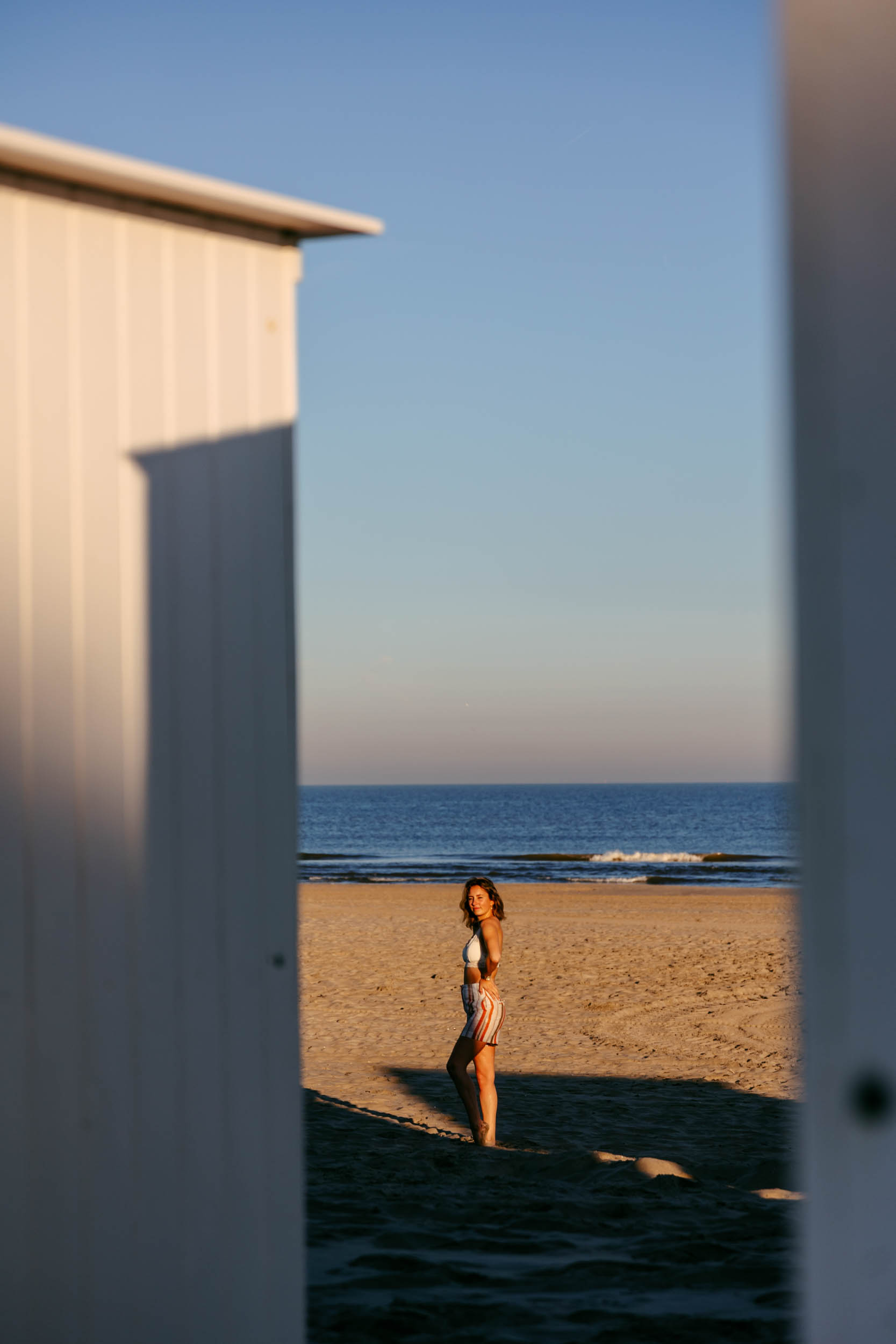 A woman poses in front of a beach hut taking beach photos.