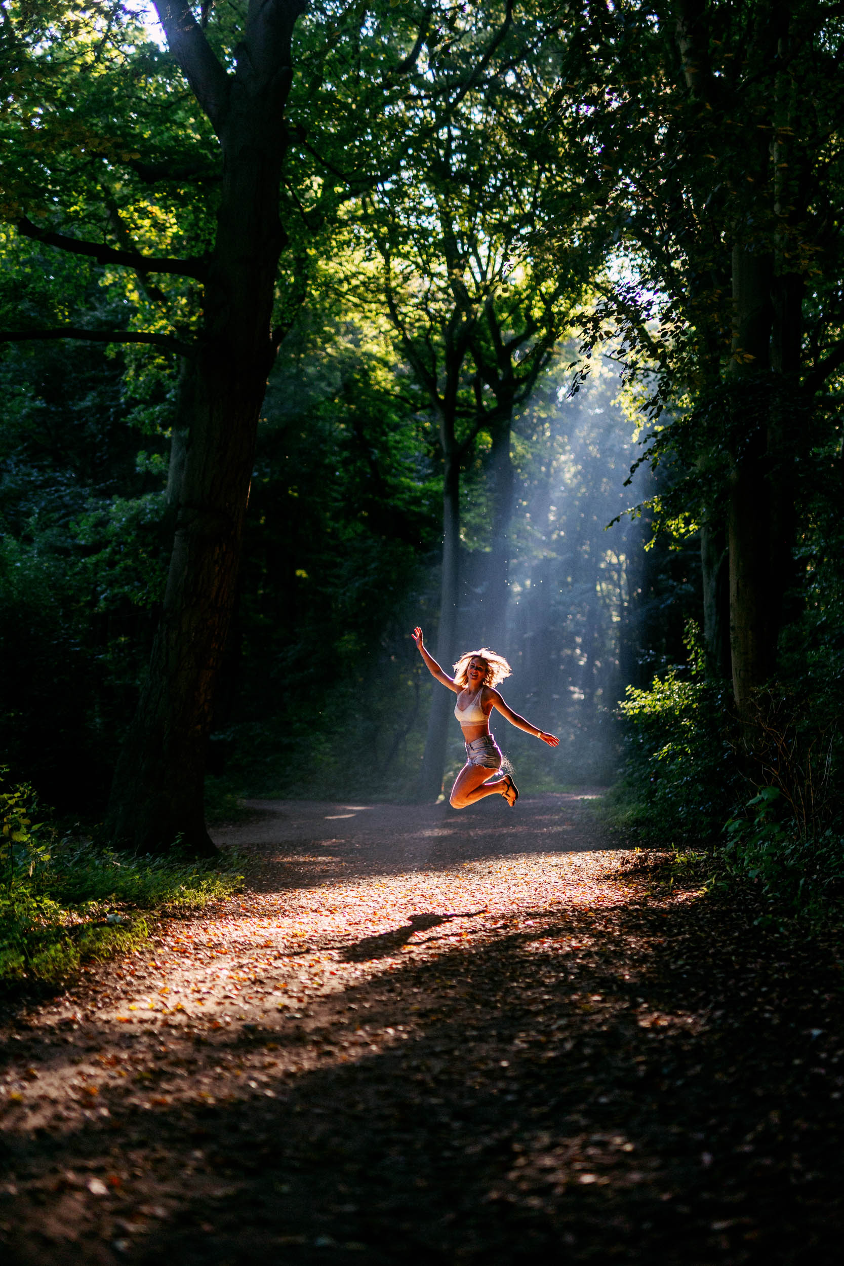 A girl leaps through the air between a strand of trees as sunlight peeks through the foliage.