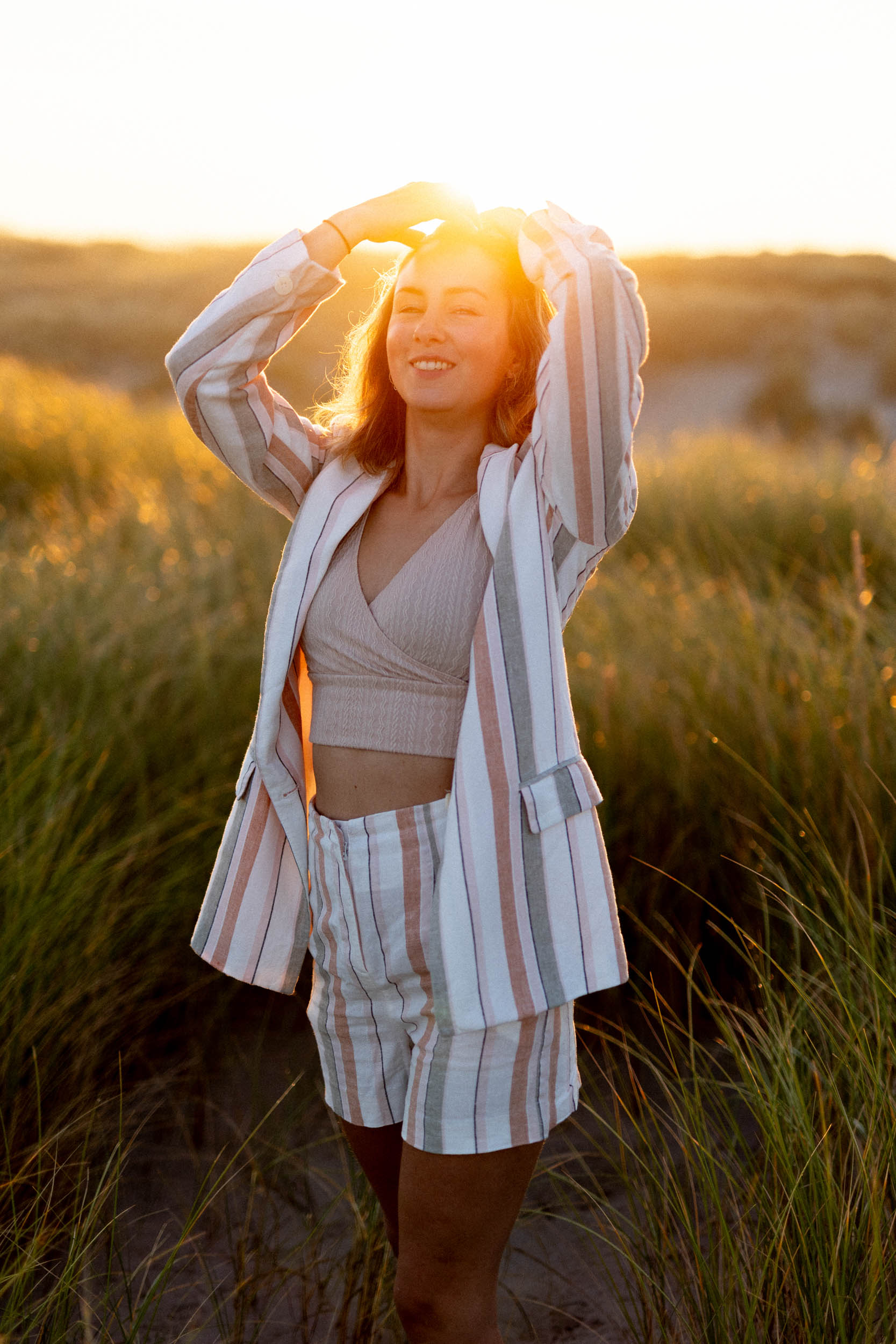 A woman in a striped blazer poses for beach photos in tall grass at sunset.