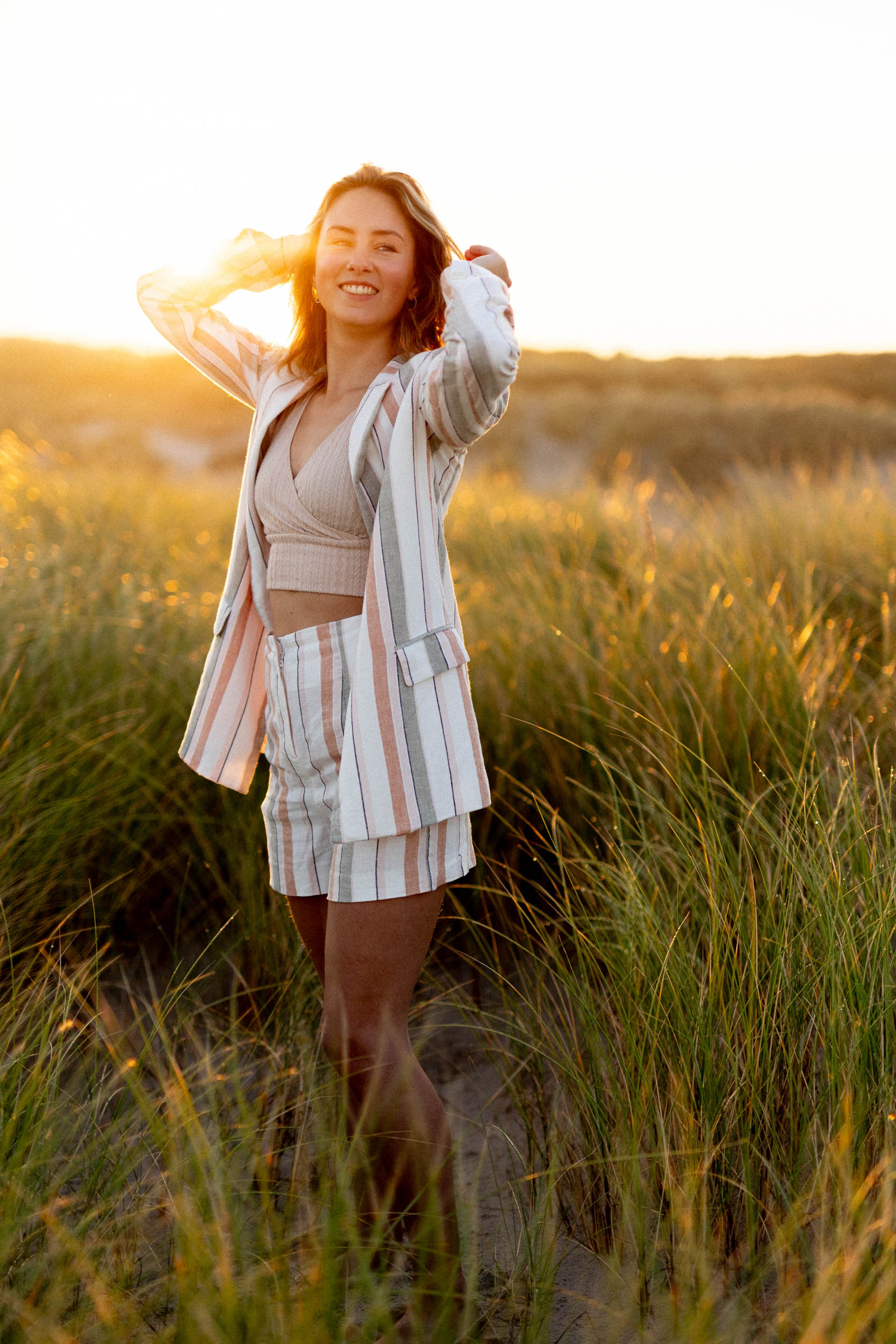 A woman in a striped blazer standing in the tall grass at sunset, captured in beach photos.