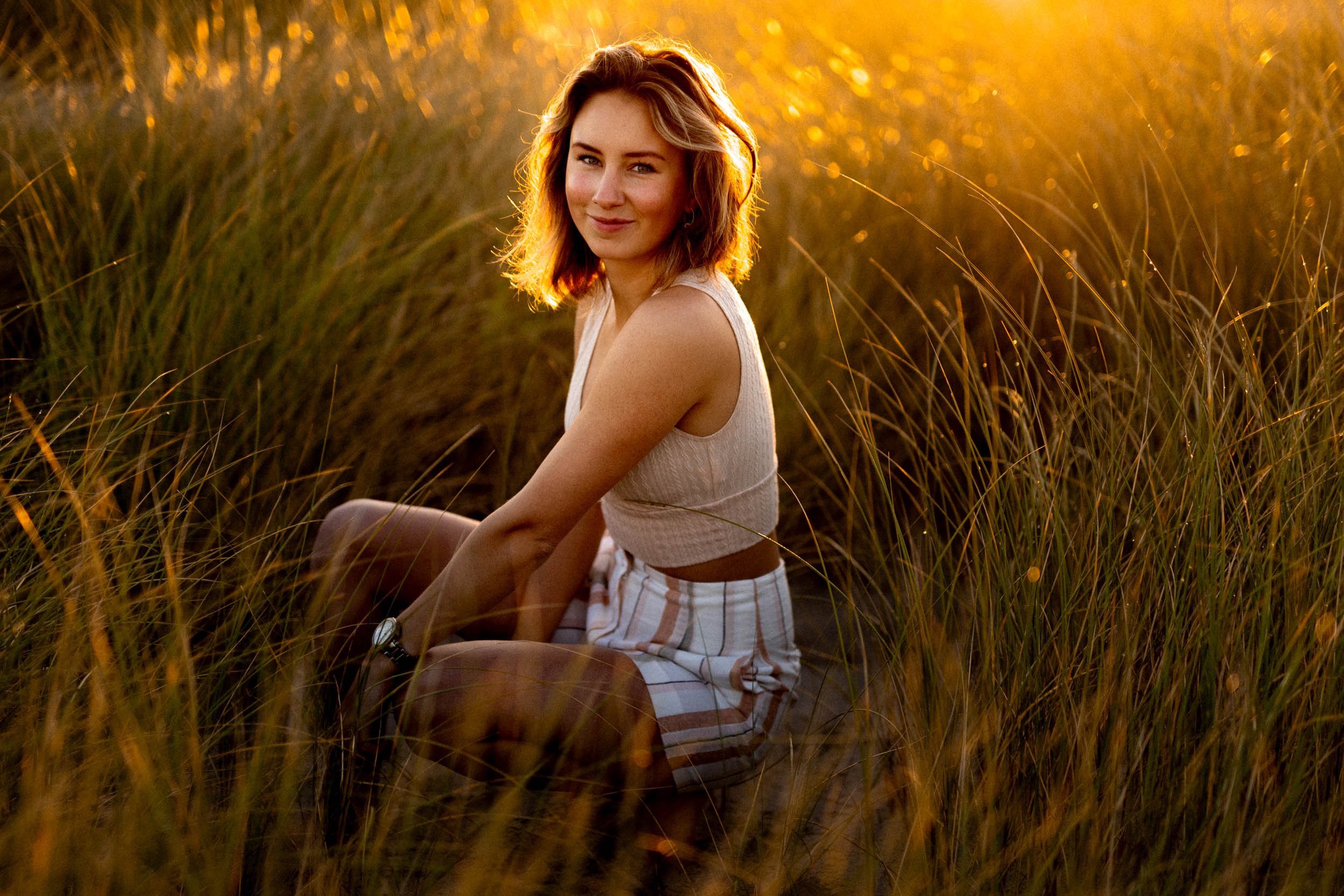 A young woman sits in tall grass at sunset, captured in a beautiful beach photo.