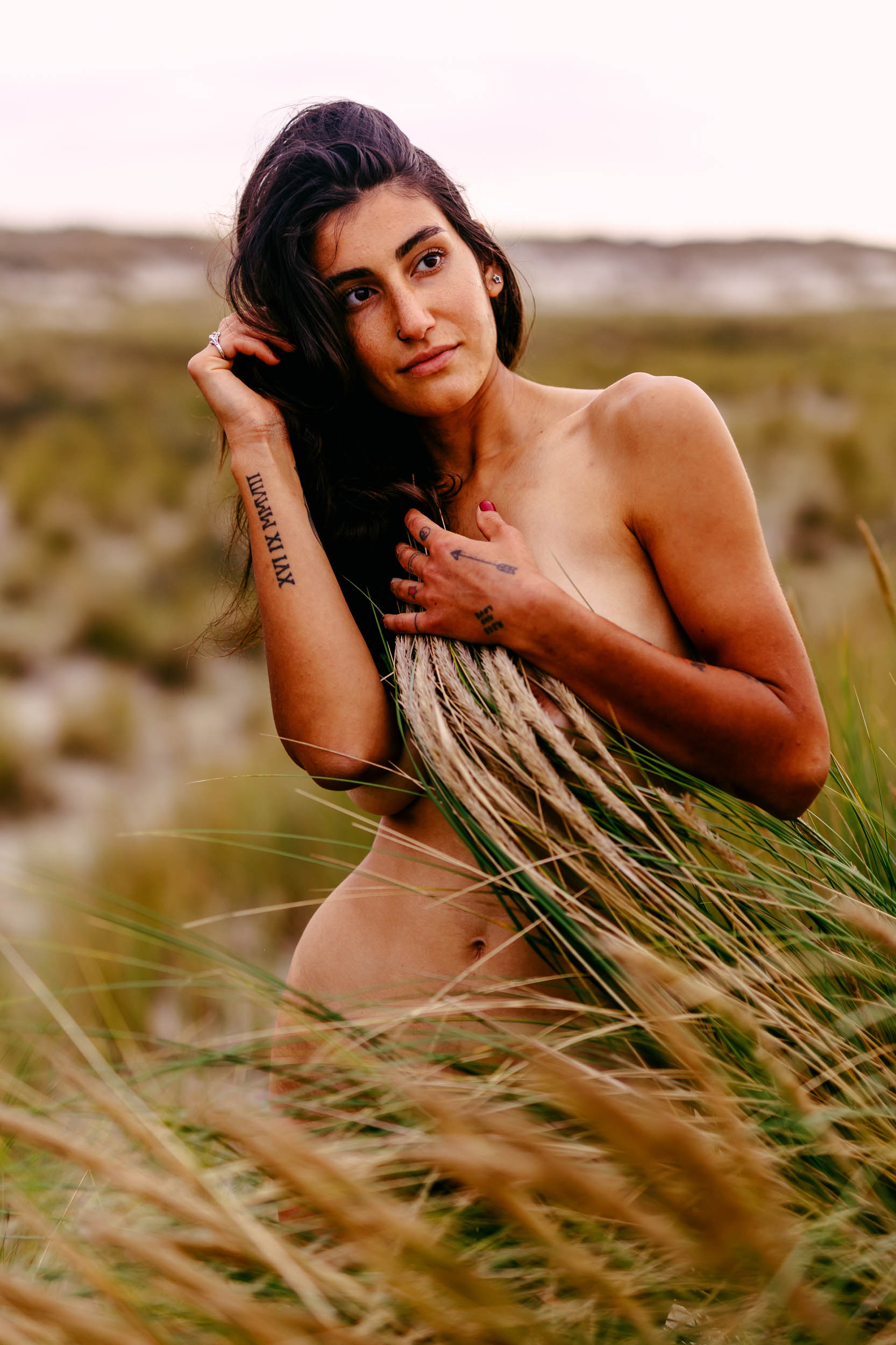 A naked woman poses strikingly in the tall grass during a captivating beach photo shoot.