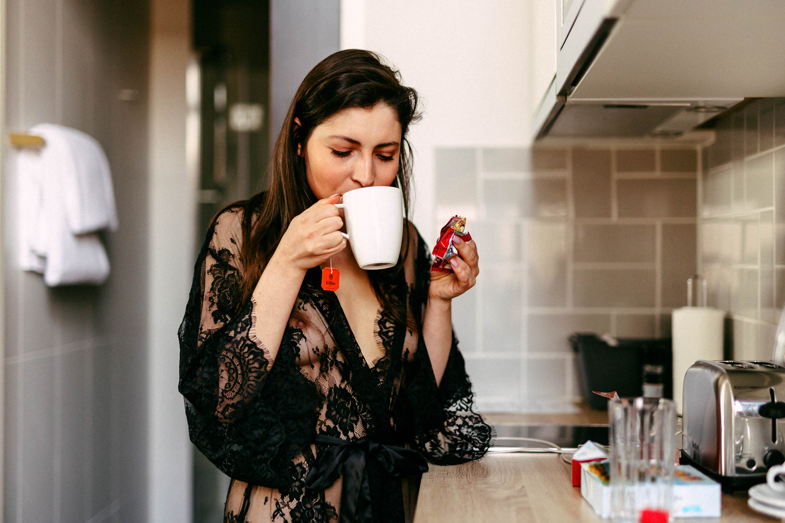 A woman in a black robe drinking coffee in a kitchen.