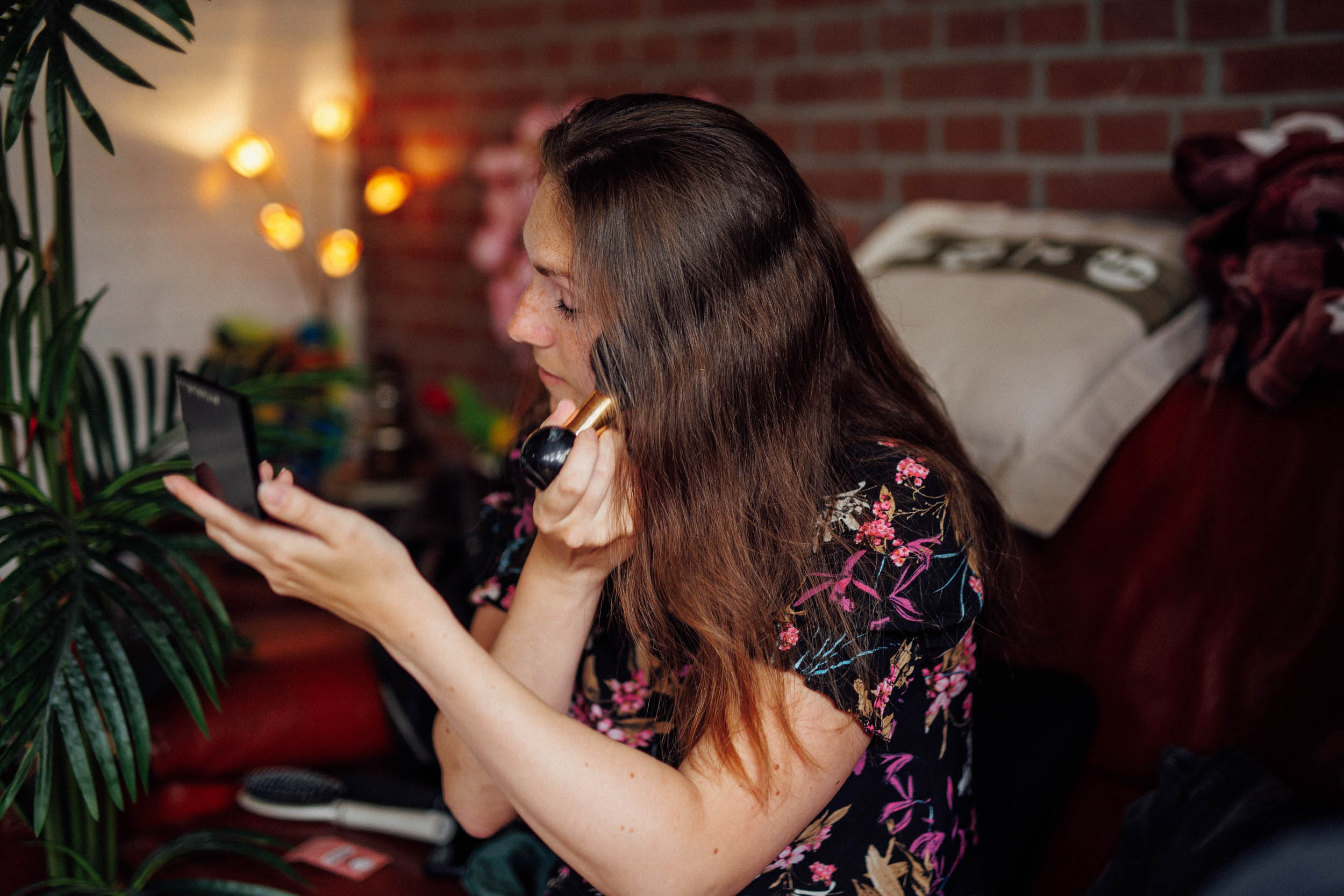 Een vrouw met lang haar brengt make-up aan terwijl ze in een handspiegel kijkt in een gezellige kamer met zachte verlichting en een plant, waardoor een intieme boudoirfotoscène ontstaat.