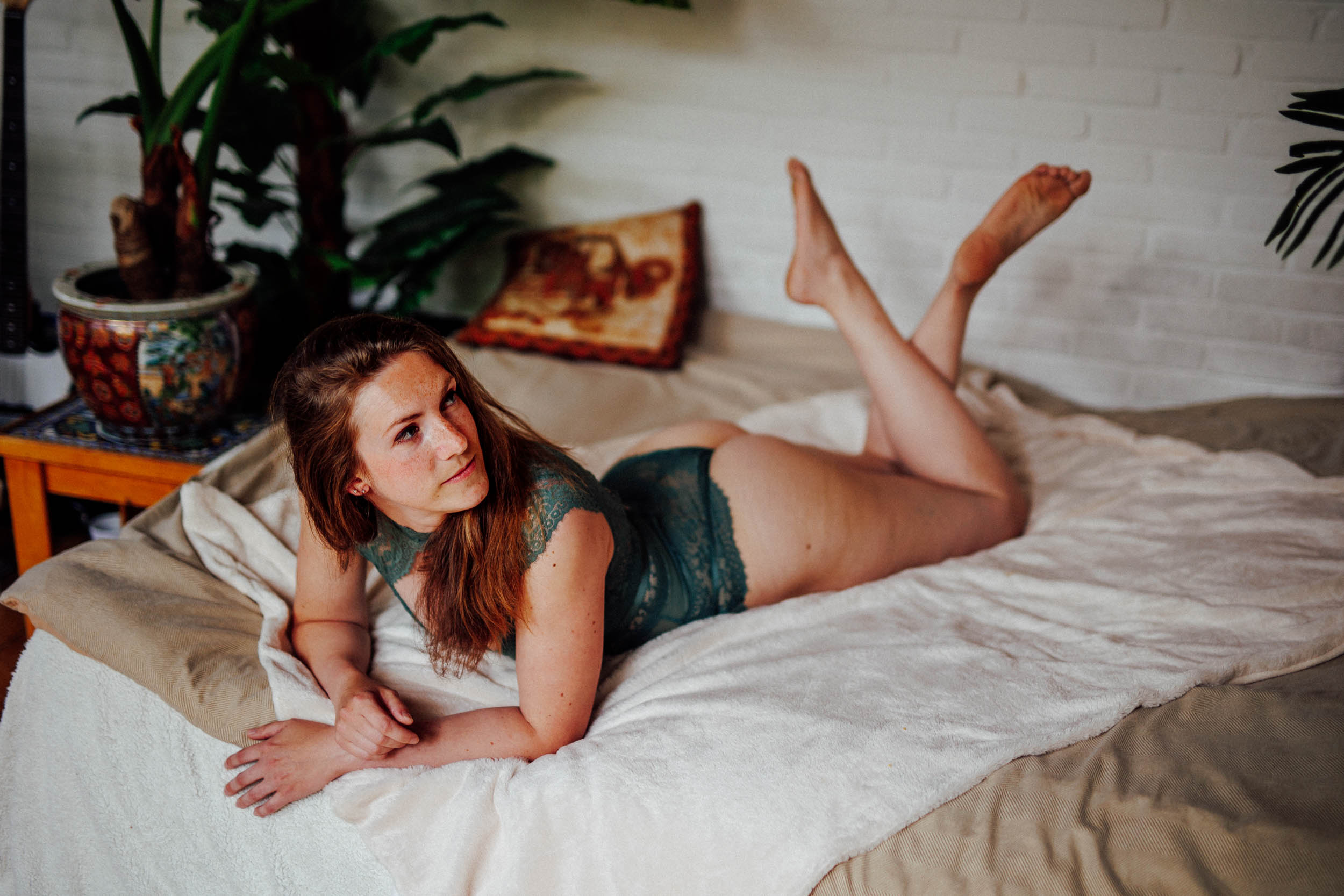 Een vrouw in een groen kanten lingeriesetje ligt op haar buik op een bed, ondersteund door kussens. De scène, perfect voor boudoirfoto's, bevat planten en een bijzettafel op de achtergrond.