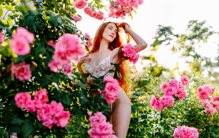 Een vrouw poseert sierlijk tussen roze rozenstruiken, wat de essentie van outdoor boudoirfotografie belichaamt. Ze draagt een bloemenoutfit en houdt haar haar zachtjes vast, terwijl de lucht erboven helder en zonnig blijft.