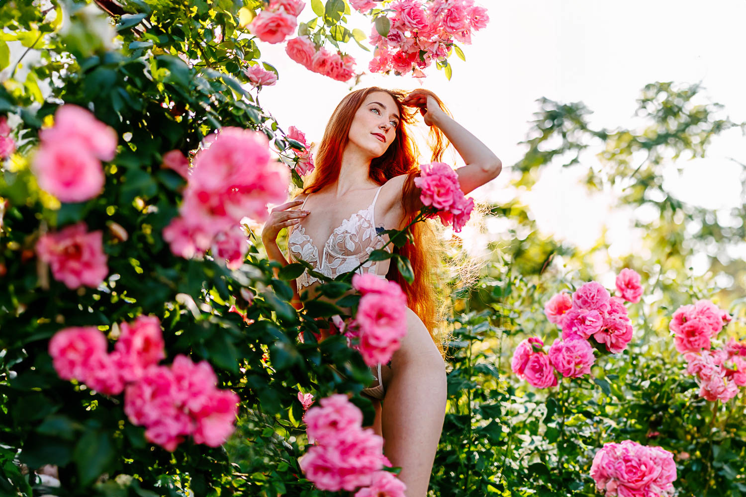 Een vrouw poseert sierlijk tussen roze rozenstruiken, wat de essentie van outdoor boudoirfotografie belichaamt. Ze draagt een bloemenoutfit en houdt haar haar zachtjes vast, terwijl de lucht erboven helder en zonnig blijft.