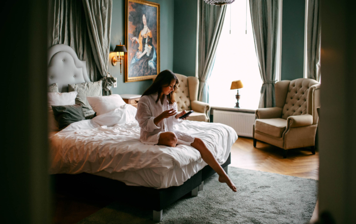 Een vrouw in een wit gewaad zit op een opgemaakt bed, met een glas en een boek in een kamer met groene muren, twee fauteuils en een groot schilderij. Daglicht stroomt naar binnen door een raam met grijze gordijnen.