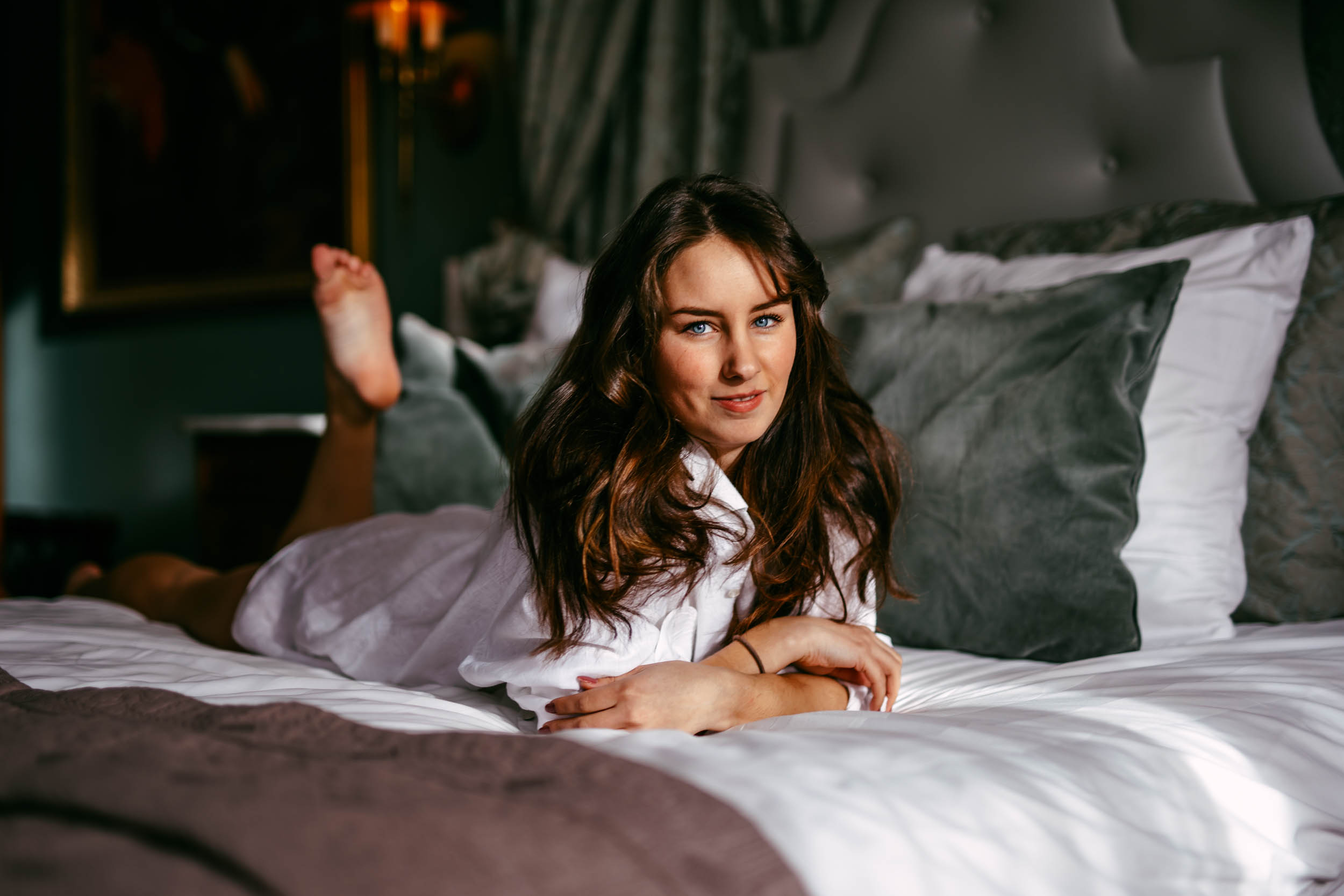 A woman with long brown hair, dressed in a white robe, lies on her stomach on a neatly made bed with grey and white pillows in a dimly lit room.