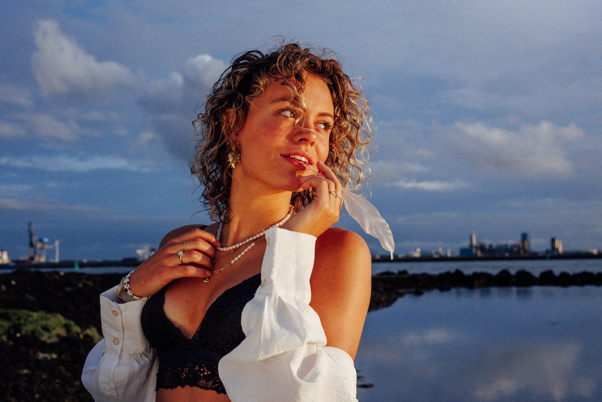 A person with curly hair stands by a body of water, dressed in a black top and white shirt. The background consists of a skyline and cloudy sky.