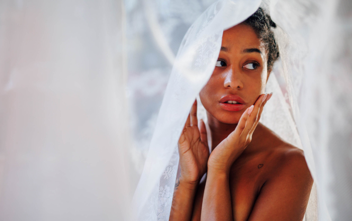 A woman with a contemplative expression is draped in a translucent white fabric, touching her face with both hands.