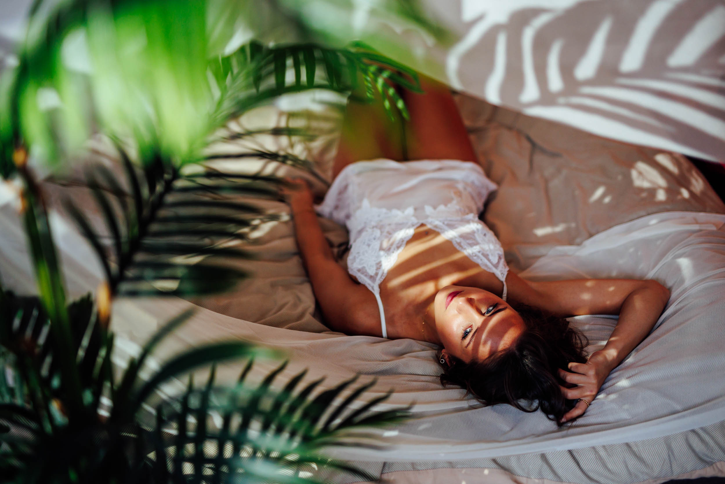 Een vrouw in een wit kanten pakje ligt op een bed, omringd door groene bladplanten en schaduwpatronen.