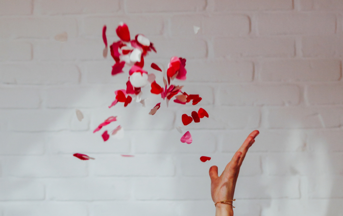 Een hand gooit rode en witte bloemblaadjes in de lucht tegen een witte bakstenen muur als achtergrond. Het is de perfecte setting voor een boudoirfotoshoot.