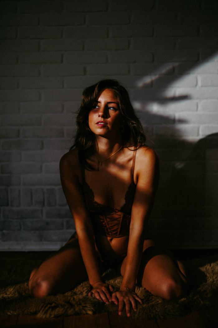 Woman in lingerie kneeling on a soft carpet in a dim room with a brick wall, the Boudoir Preset Pack casts shadows and golden sunlight on her face and body.