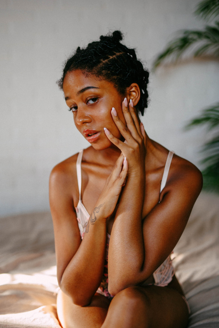 A woman poses indoors, bathed in golden sunlight, dressed in a light-coloured top. With one hand on her face and the other resting on her chest, she sits gracefully on a bed with plants in the background. Her short braided hair captures the essence of the Boudoir Preset Pack perfectly.