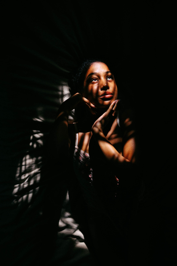 A person sits with his chin on his hands, illuminated by a pattern of light shining through blinds in an otherwise dark room. It is reminiscent of the warm glow of golden sunlight from a Boudoir Preset Pack.