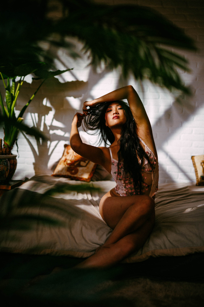 A woman with long dark hair poses on a sofa, framed by golden sunlight and shadows of plants, dressed in a floral outfit. A white brick wall and potted plants serve as the backdrop, capturing the essence of a Boudoir Preset Pack.