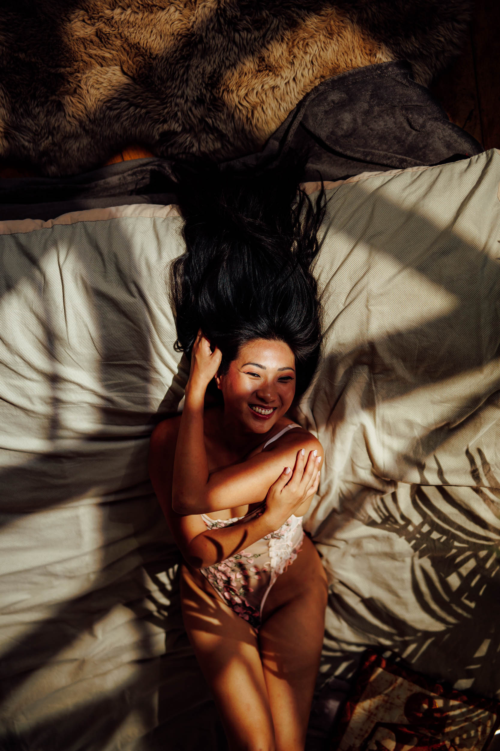 A woman with long dark hair lies on her back on a bed, smiling, with her arms crossed over her chest and partially covered in sunlight; the scene produces beautiful photographs of an Asian woman.