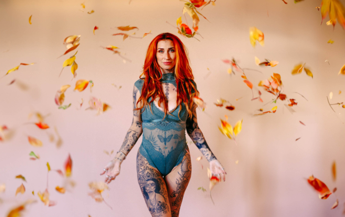 Een vrouw met lang rood haar en tatoeages staat in een blauwgroen kanten bodysuit terwijl de herfstbladeren om haar heen vallen.